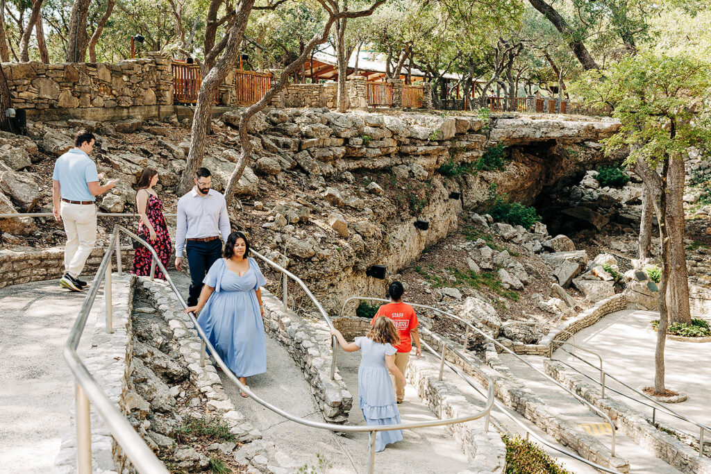 People walk along a winding path with railings through a rocky, tree-filled park area near the natural bridge caverns. Some wear summer clothing, and the landscape features stone walls and a cave-like entrance in the background.