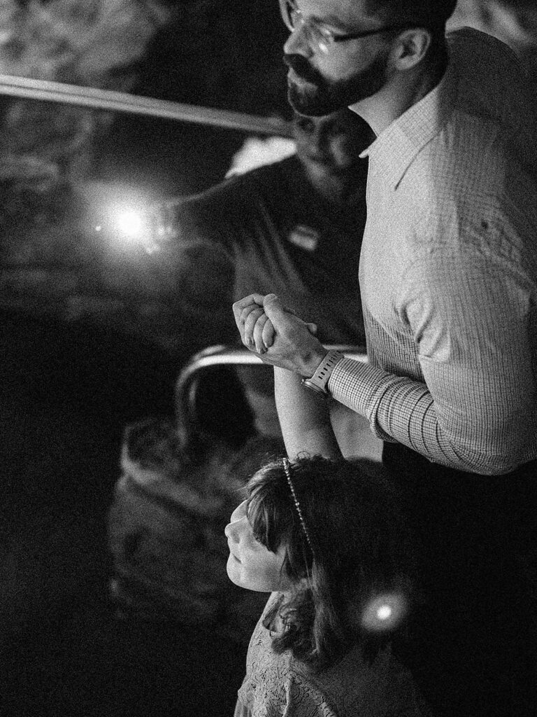 A black-and-white photo of a man and a young girl holding hands, looking upward—perhaps at the vast ceilings of Natural Bridge Caverns. The girl wears a headband; the bearded, bespectacled man stands beside her. Another person in the background holds a bright light.