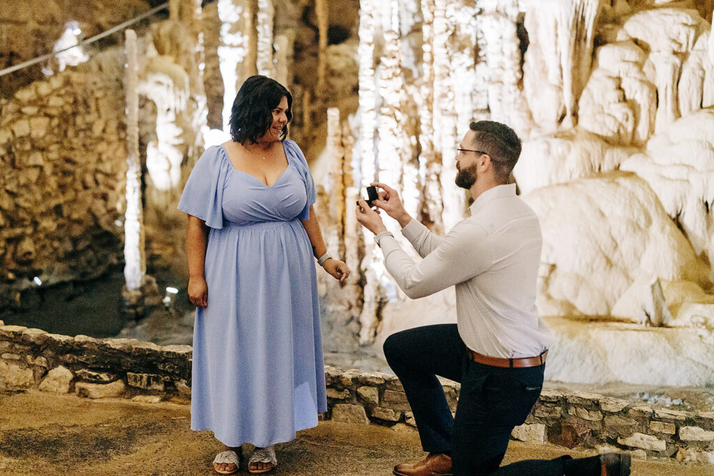 A man kneels to propose with an open ring box inside the awe-inspiring Natural Bridge Caverns, as a woman in a light blue dress smiles in surprise and happiness among stunning rock formations.