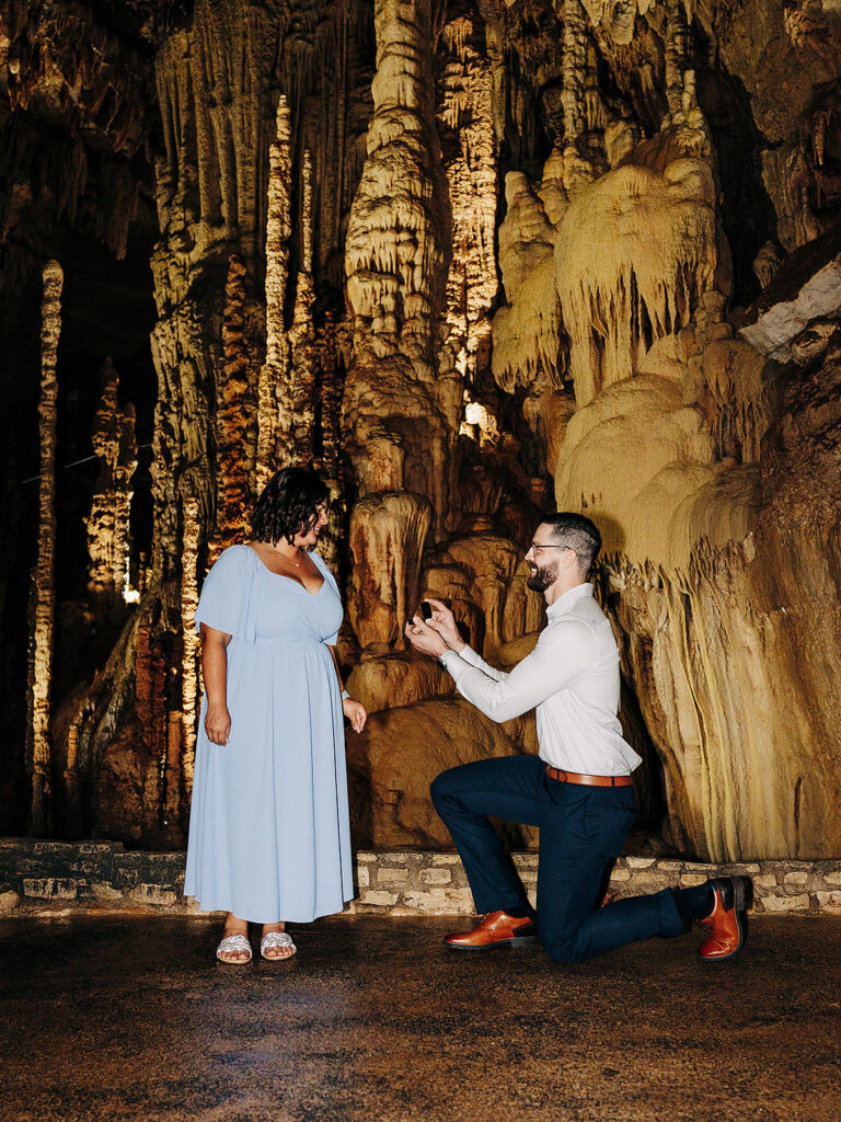 A man kneels and makes a heartfelt proposal to a woman in a blue dress inside Natural Bridge Caverns, surrounded by dramatic stalactites and stalagmites illuminated by warm lighting.