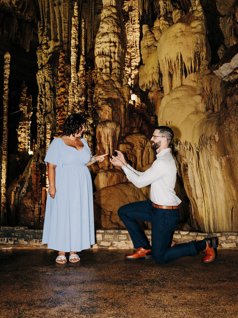 A man kneels and proposes with a ring to a woman in a light blue dress inside the awe-inspiring Natural Bridge Caverns, surrounded by dramatic rock formations and shimmering stalactites.