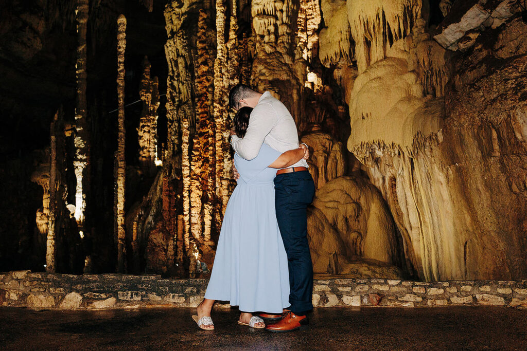 A couple embraces closely in Natural Bridge Caverns, surrounded by tall, illuminated stalagmites and stalactites—an intimate moment that feels perfect for a proposal. The woman wears a light blue dress; the man is in a white shirt and blue pants.