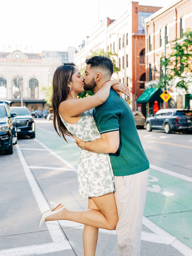 A couple embraces and kisses in the middle of a Denver city street on a sunny day. The woman lifts one leg as she hugs the man, with brick buildings and the historic Millenium Bridge visible in the background.