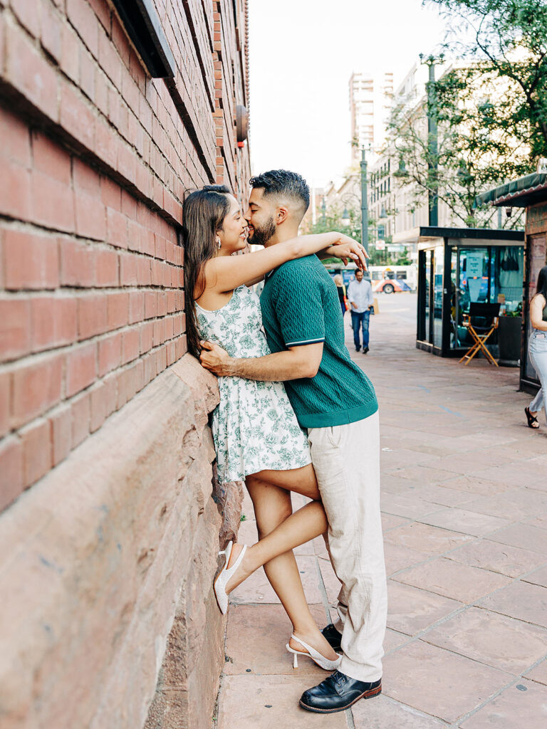 A couple embraces and smiles at each other while standing close to a brick wall on a city sidewalk near Denver&rsquo;s Millennium Bridge. The woman wears a floral dress and heels, and the man wears a green shirt as people and buildings bustle in the background.