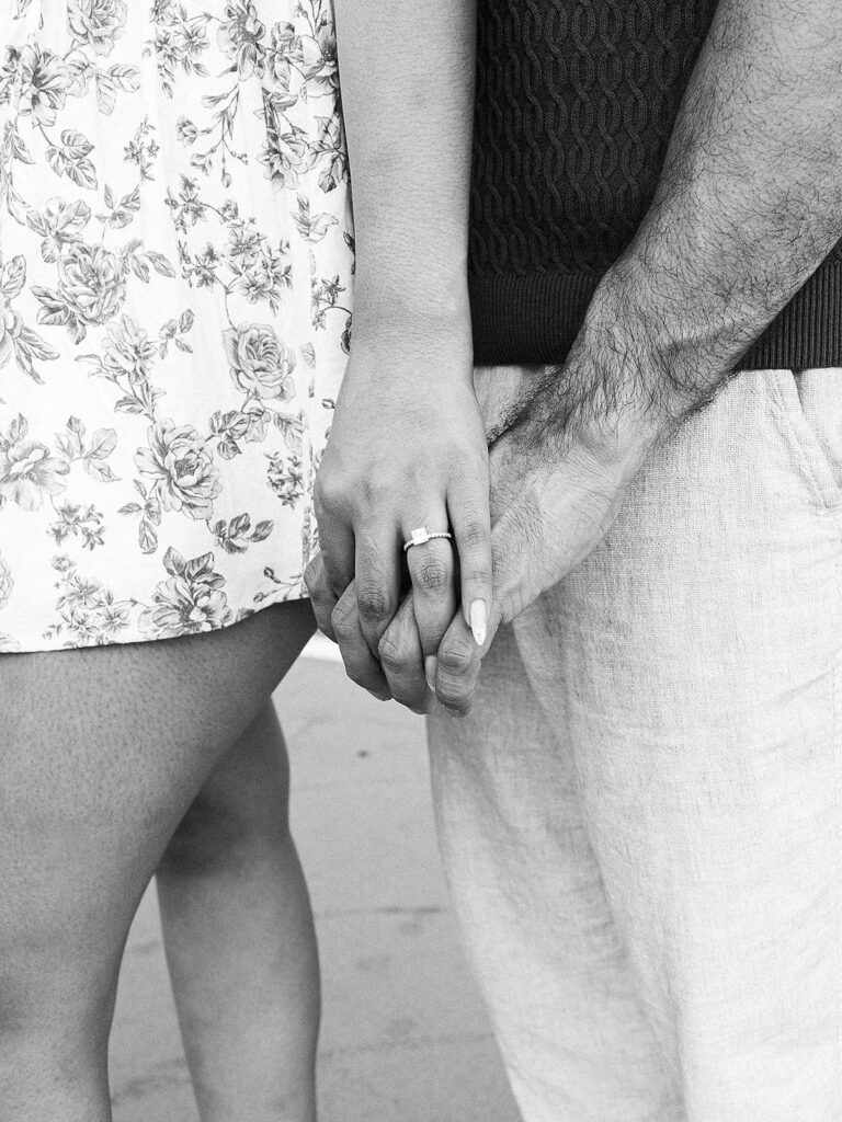 A black-and-white photo of a couple holding hands on the Millenium Bridge in Denver. The woman's floral dress and engagement ring are visible, while the man's hand rests gently over hers, their closeness suggesting intimacy and affection.
