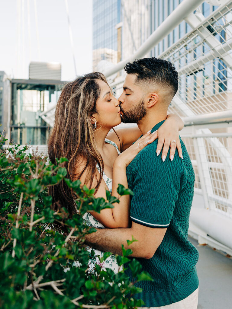 A couple kisses passionately on Denver's Millennium Bridge, with greenery in the foreground and tall buildings in the background. The woman has long hair and wears a light dress; the man wears a green textured shirt.