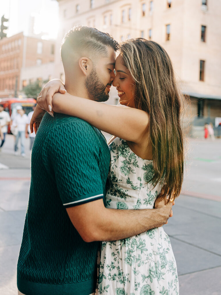 A couple embraces closely on a city street near Denver&rsquo;s Millennium Bridge, gazing into each other's eyes and smiling. The woman wears a white floral dress, and the man is in a green shirt. Sunlight glows behind them, with blurred buildings and people in the background.