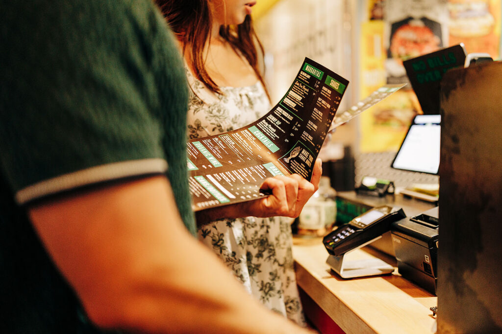 A woman holding a menu stands at a counter, appearing to place an order in a Denver restaurant. Another person is beside her, and a card payment terminal is visible on the counter.