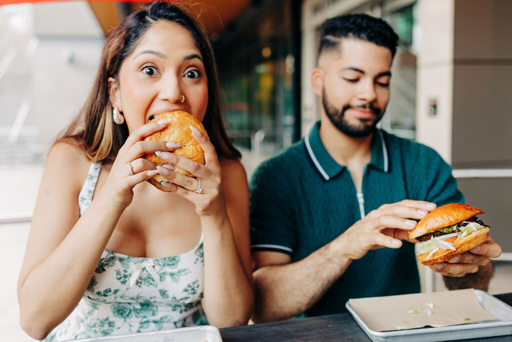 A woman eagerly takes a big bite of a burger while a man next to her smiles and prepares to eat his own at an outdoor restaurant table near Denver's iconic Millennium Bridge.