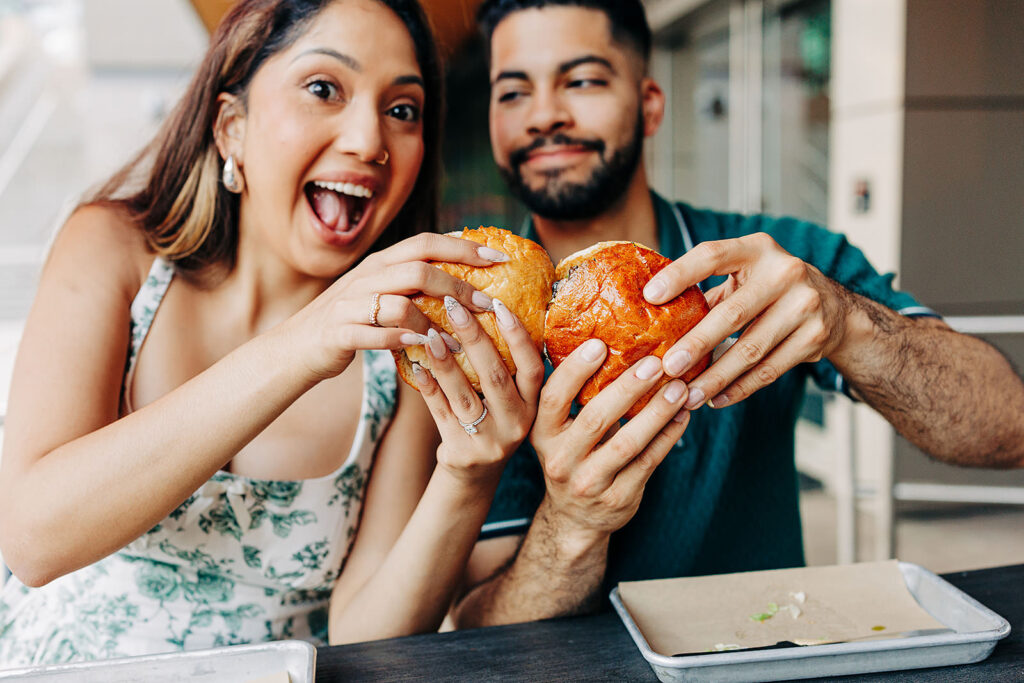 A smiling woman and man sit together at a table in Denver, holding up large burgers with both hands as if to toast. The woman looks excited with her mouth open and the man smiles gently. Empty trays are in front of them.