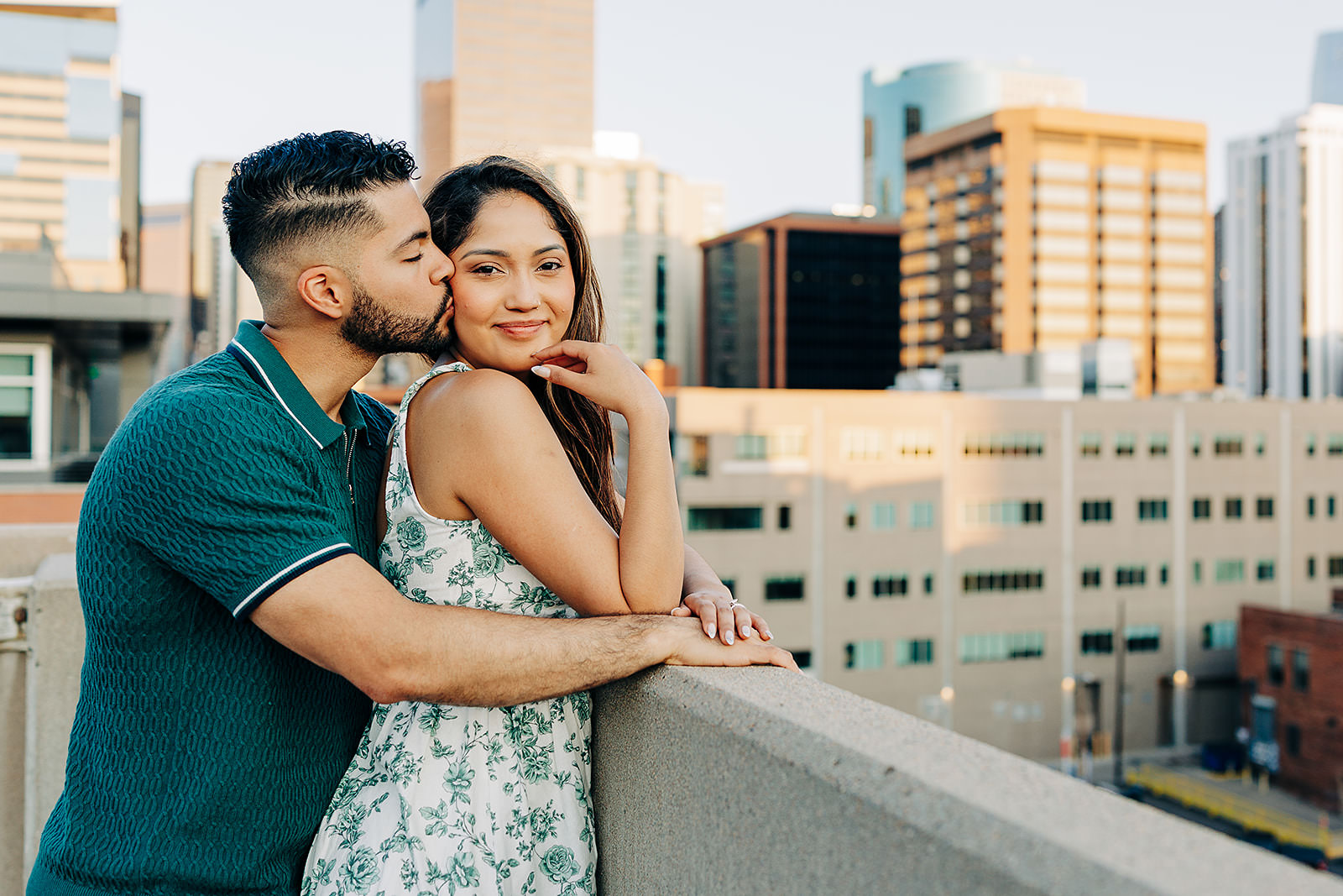A man kisses a woman on the cheek as they stand close together on a rooftop in Denver, with tall buildings and the cityscape in the background. The woman smiles, resting her hand on the man's chest.