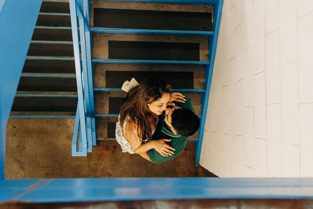 A couple embraces on the Millenium Bridge staircase in Denver, viewed from above. The woman in a white dress smiles at the camera while the man in a green shirt holds her. Blue railings and grey steps frame the scene.