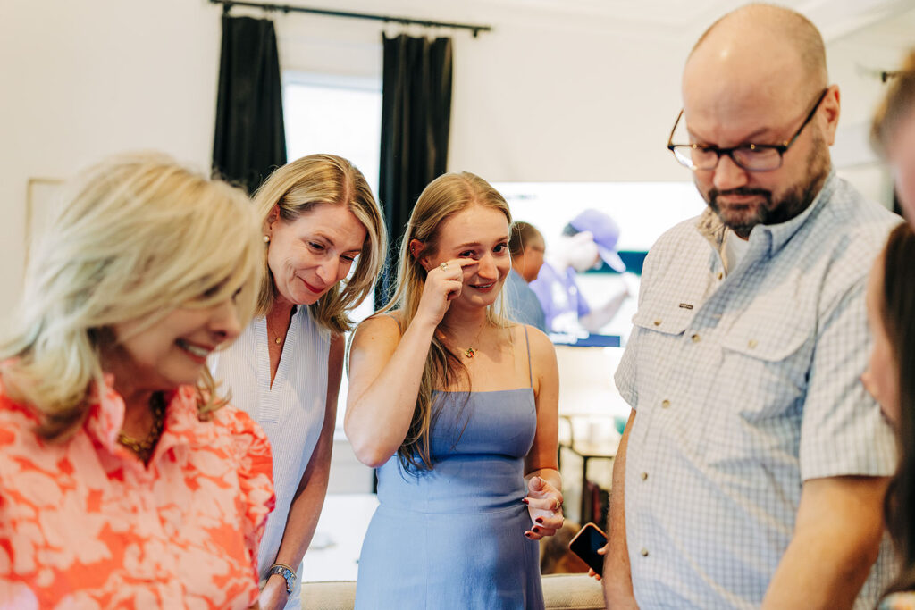 A group of five adults, smiling and engaged in conversation, stand together indoors&mdash;perhaps celebrating a recent proposal in the King William District. One woman in a blue dress wipes her eye as others look on warmly. A TV and dark curtains are visible behind them.