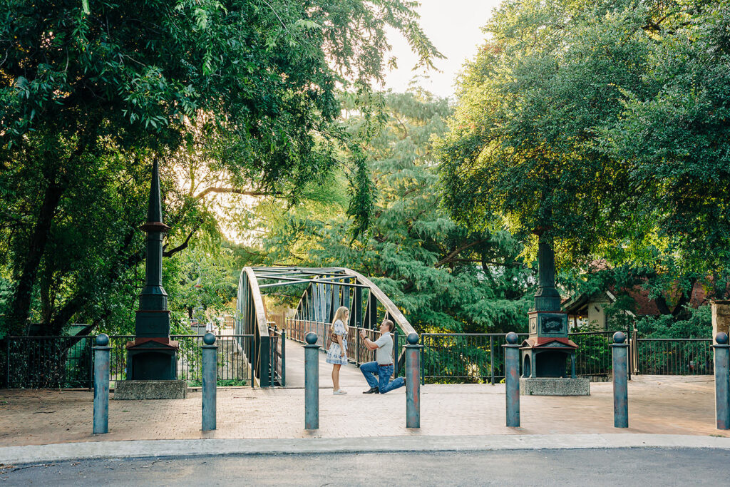 A man kneels in proposal to a woman at the entrance of a tree-lined pedestrian bridge on a sunny day. The peaceful scene, with lush greenery and soft sunlight, highlights why it&rsquo;s among top San Antonio Proposal Locations.
