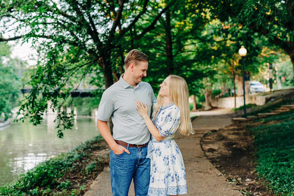 A couple stands on a tree-lined path by a river, smiling at each other. The woman wears a light blue floral dress, the man a gray shirt and jeans. It's a bright day&mdash;one of the perfect San Antonio proposal locations surrounded by lush greenery.