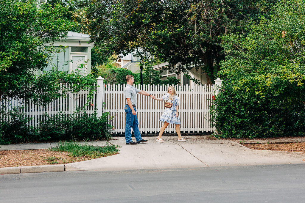 A man and a woman stand on a sidewalk in front of a white picket fence and lush greenery&mdash;one of the picturesque San Antonio proposal locations. The man gently reaches out to touch the woman&rsquo;s arm as she walks past, looking back at him.
