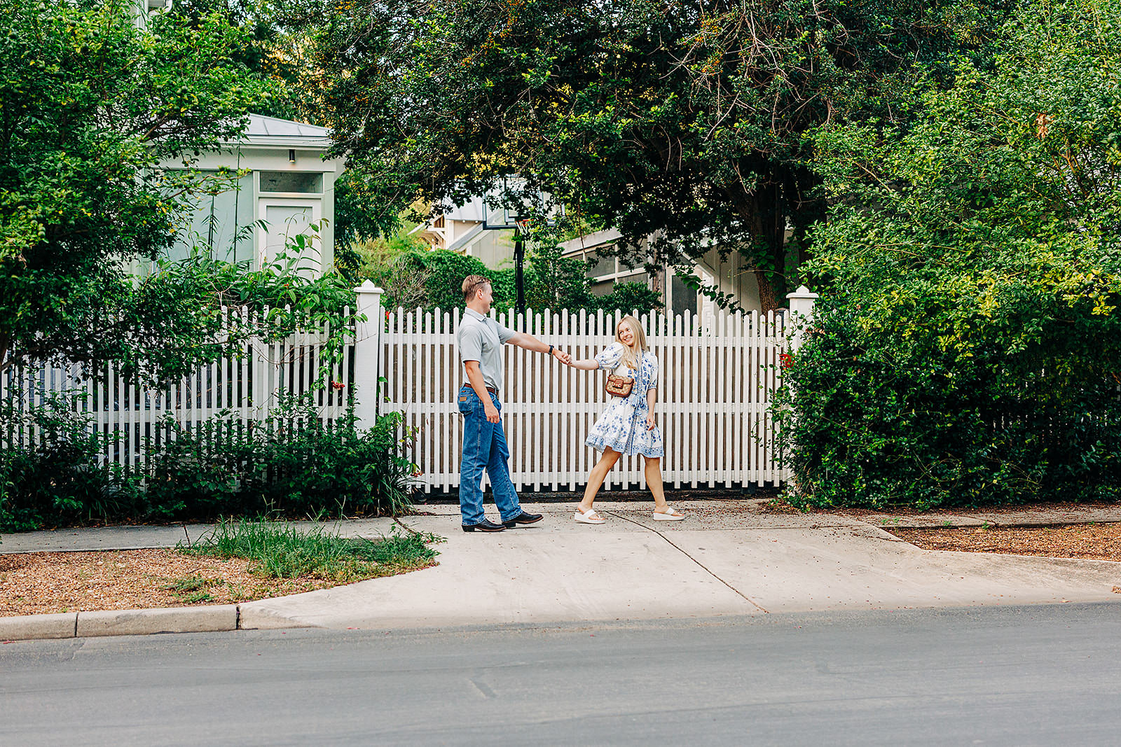 A man and a woman holding hands walk past a white picket fence surrounded by greenery in the King William District on a sunny day. The woman wears a light dress and sandals, while the man wears jeans and a t-shirt, hinting at an intimate proposal.