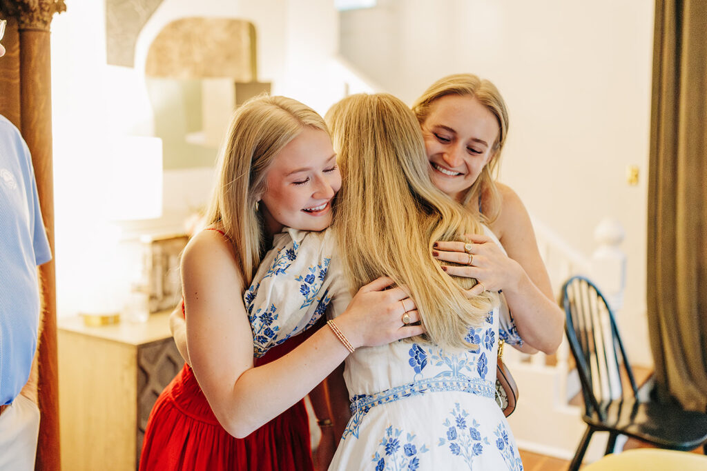 Three young women with blonde hair share a warm group hug and smile in a bright, cozy indoor setting in the King William District. Two wear white and blue dresses, one a red skirt, expressing joy&mdash;perhaps celebrating an exciting proposal.