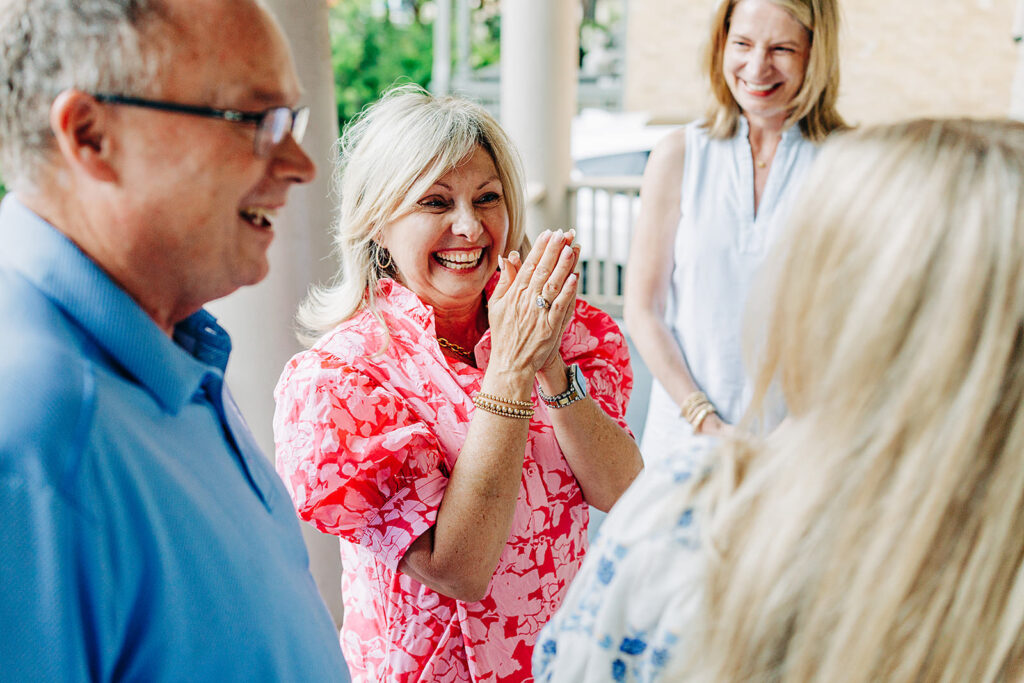 A woman in a pink floral blouse smiles and claps her hands joyfully while discussing a proposal with three others on a porch in the King William District. Everyone appears happy and engaged in conversation.