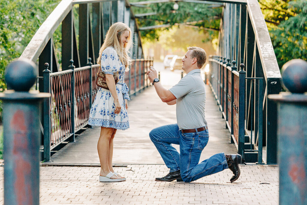 A man kneels on one knee, holding out a ring in a heartfelt proposal to a surprised woman on a bridge in the lush King William District.