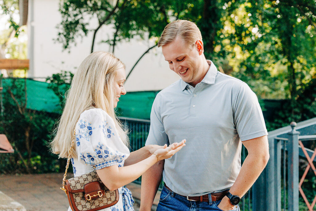 A woman and a man stand outdoors in the King William District, smiling and looking at the woman's hand&mdash;perhaps after a proposal. She wears a white dress with blue patterns, while he is in a light gray polo shirt and jeans. Green trees and a fence are behind them.