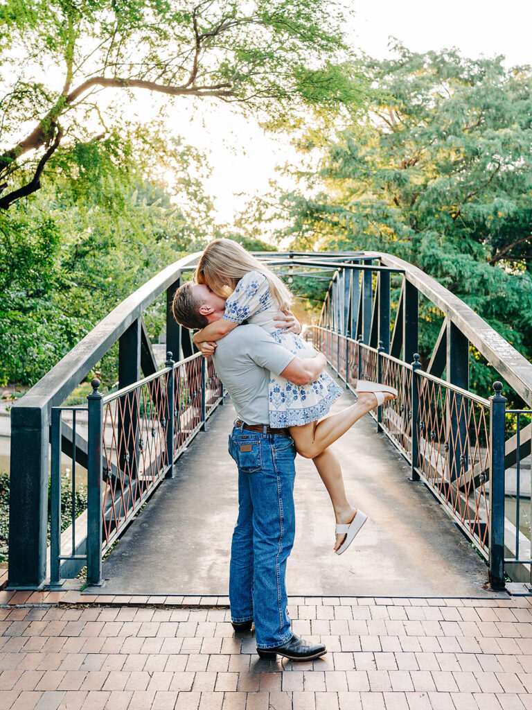 A man stands on a bridge in the King William District, lifting a woman in a joyful embrace after a proposal. She wraps her arms and legs around him, both smiling as trees and sunlight frame the moment.