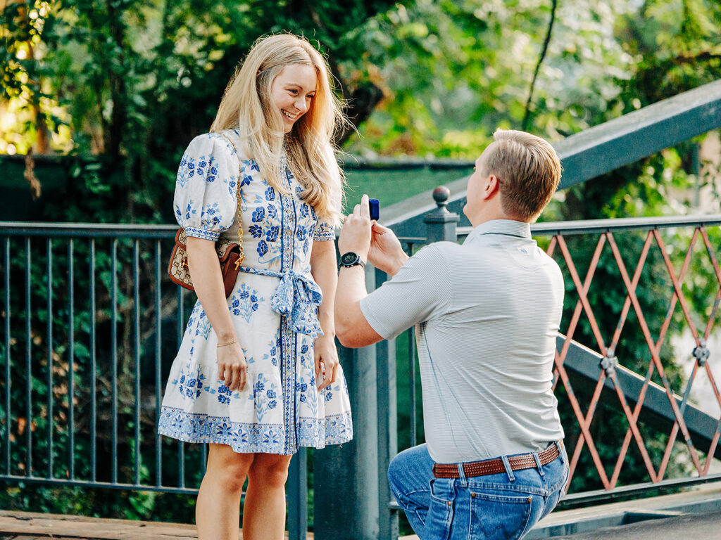 A man kneels on one knee, holding an open ring box in a heartfelt proposal to a smiling woman in a white and blue dress. They are outdoors on a wooden bridge in the scenic King William District, surrounded by lush greenery.
