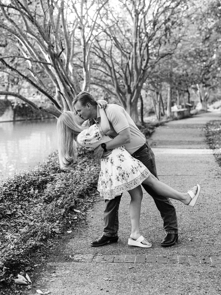 A man dips and kisses a woman on a tree-lined riverside path in the King William District. The woman wears a floral dress; the man, jeans and a polo shirt. This romantic, serene moment feels like a proposal, captured in black and white.