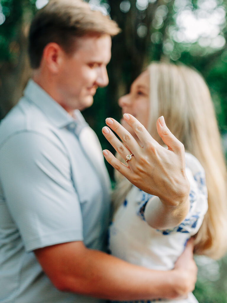 A couple embraces outdoors in the King William District, the woman proudly displaying her engagement ring after a heartfelt proposal. The softly blurred greenery provides a romantic backdrop.